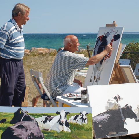 Malevent am Meer, Hohwachter Bucht, Maler sitzt auf Stuhö, vor ich eine Staffelei mit Leinwand, Maler malt Kuh, neben dem Maler Tisch mit Pinsel und Farben, Zuschauer hinter dem Maler, im Hintergrund die Ostsee, im Vordergrund weitere Leinwände mit Kuhmot