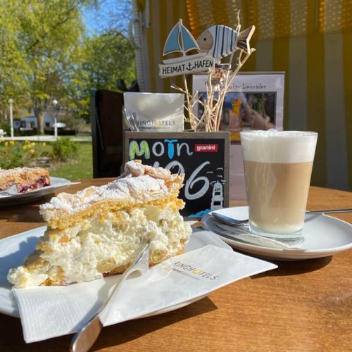 Nahaufnahme gedeckter Tisch im Kurparkcafé, Sitzmöglichkeit mit Strandkorb, auf dem Tisch ein Teller mit einem Stück Sahnetorte und ein Teller mit Obststreuselkuchen, Latte macchiato, Aufsteller Tischnummer, Aufsteller hausgemachte Limonaden