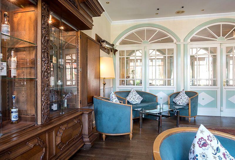 Whiskeylounge Hotel Hohe Wacht, links großer Schrank mit Schnitzereien und Glasfronten, im Schrank verschiedenste Sorten Whiskey, gemütliche Sitzecke mit Sesseln sowie Glastisch, im Hintergrund Fenster, Blick in den Flur