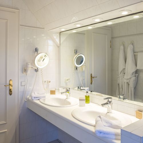 2-Zimmer-Komfortappartement Bad mit zwei Waschbecken, Spiegelfront, Kosmetikspiegel, Föhn, Handtücher, Kosmetikproben, im Spiegel spiegeln sich zwei Bademäntel hängend am Haken
