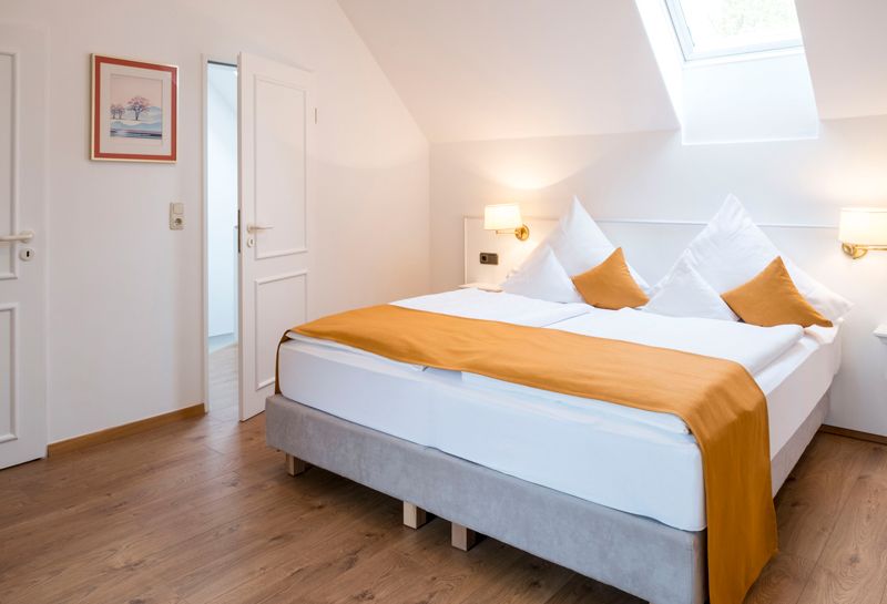 Schlafzimmer mit Boxspringbett unter einer Schräge, in der Dachschräge Fenster, links und rechts vom Bett Lampen und Ablageflächen, an der Wand hängt ein Bild, zwei Türen