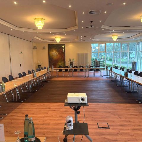 Tagungsraum im Hotel Hohe Wacht, Tische in Stühle in U-Form aufgestellt, eingedeckt mit Gläsern, Getränken und Schreibblöcken, an der Decke Kronleuchter, rechts helle Fensterfront, im Vordergrund Beamer, links Trennwand zum anderen Raum