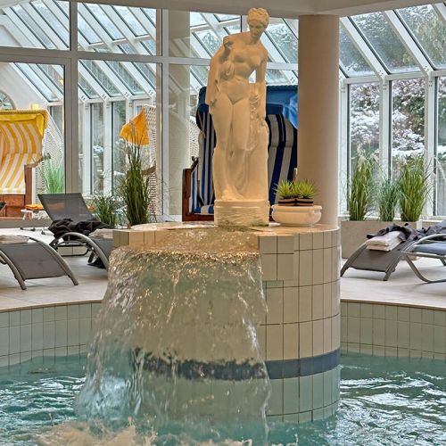 Wellness und Spa Hohe Wacht, Schwimmbereich mit Schwimmbecken, Statue mit Wasserfall, im Hintergrund Ruhebereich mit Strandkörben und Liegen