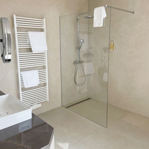 Bad Kurpark-Suite, links Waschtisch mit Waschbecken und Kosmetikspiegel, an der Wand daneben Handtuchheizkörper, Dusche mit Glaswand, durch die Glaswand sieht man die Duscharmatur