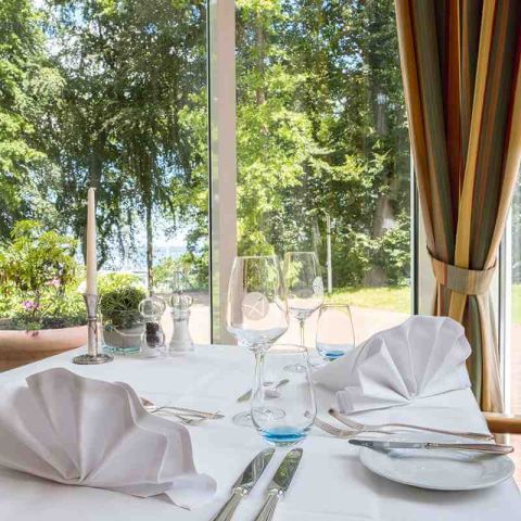 Nahaufnahme gedeckter Tisch im Parkrestaurant am Fenster, mit Teller, Besteck, Wasser- und Weingläser, Kerze, Salz-und Pfefferstreuer, gefaltete Servietten, Blick in den Kurpark auf die Ostsee