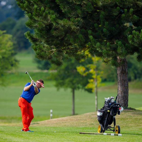 Golfen Hotel Hohe Wacht, Golfplatz, Golfer mit roter Hose schlägt Ball, auf dem Platz Bäume, rechts vom Golfer Golfbag mit Schlägern