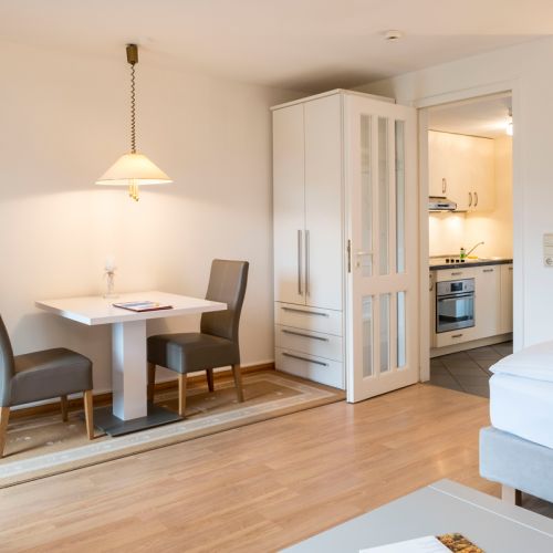 1-Zimmer-Ferienwohnung Essbereich Küche, eine Tisch mit zwei Stühlen, darunter Teppich, daneben Schrank an der Wand, rechts Eingang zur Küche mit Küchenzeile