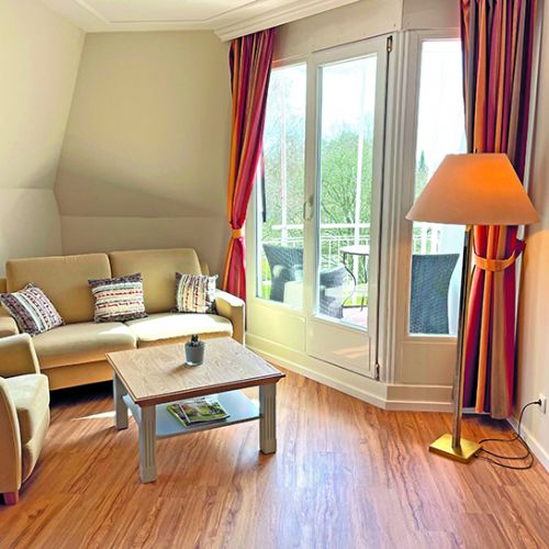 links Sitzecke mit Sofa, Tisch und Sessel, daneben Ausgang zur Loggia, auf der Loggia Sitzmöglichkeiten sowie ein kleiner Tisch, rechts neben dem Ausgang zur Loggia steht eine Stehlampe