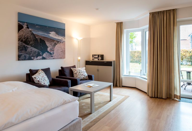 1-Zimmer Ferienwohnung Wohn- und Schlafbereich mit Boxspringbett und Sitzecke, zwei Sessel ein Tisch und Stehlampe , Sideboard mit Radio, Fensterfront, an der Wand Leinwand mit Strandmotiv