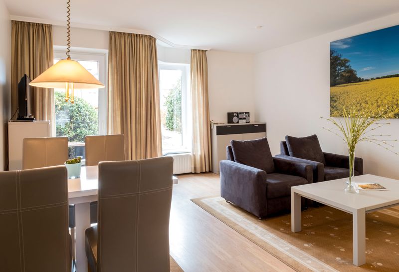 links Esstisch mit vier Stühlen, dahinter Sideboard mit TV-Gerät, rechts Sitzecke mit Tisch und 4 Sesseln, daneben an der Wand Leinwand mit Rapsmotiv, mit Blick auf die Fensterfront