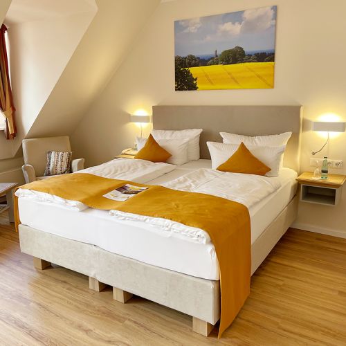 Zimmer mit Doppelbett, am Kopfende Leinwand mit Rapsmotiv, daneben Lampe an der Wand, links vom Bett am Fenster kleine Sitzecke mit Sessel und Tisch