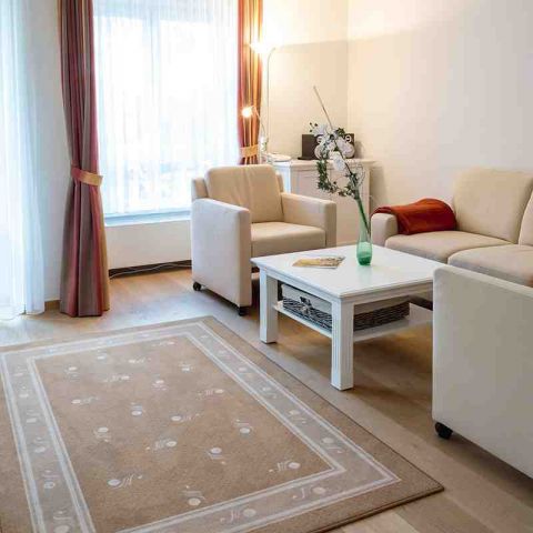 offener Wohnbereich in einer Ferienwohnung mit Sitzecke, Sofa und zwei Sessel sowie Tisch, davor Teppich, Stehlampe in der Ecke am Fenster