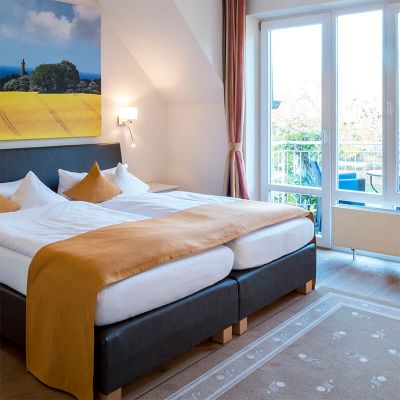 Schlafbereich im 1-Zimmer-Komfortappartement mit Schlafzimmer, an der Wand Leinwand mit Rapsmotiv, links vom Bett Terrassentür zum Balkon mit Sitzmöglichkeiten und kleinem Tisch, vor dem Bett liegt ein Teppich