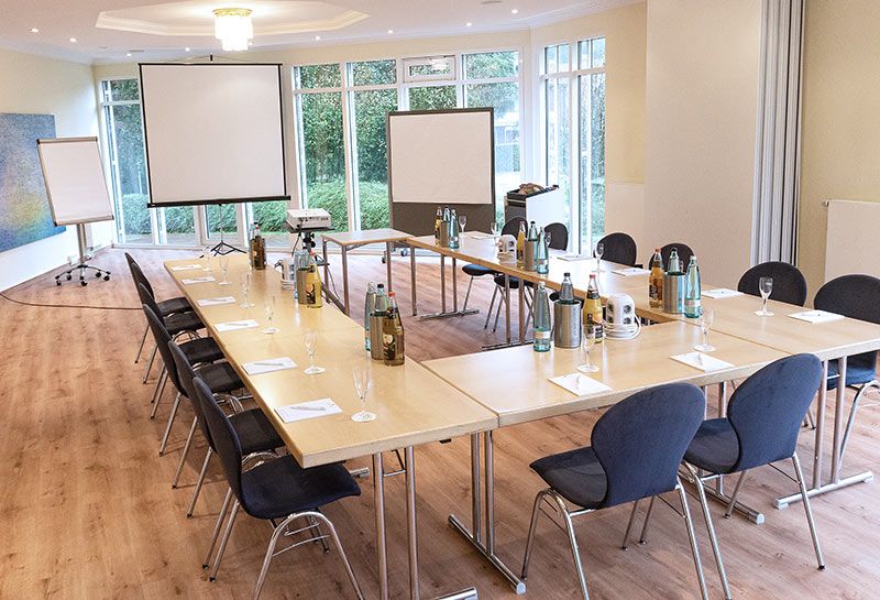 Tagungsraum Lütjenburg (Kletkamp und Neudorf) im Hotel Hohe Wacht, Bestuhlung in U-Form, eingedeckt mit Gläsern, Getränken, Schreibblöcken, im Hintergrund Beamer, Leinwand, Flipchart, Pinwand mit Papier