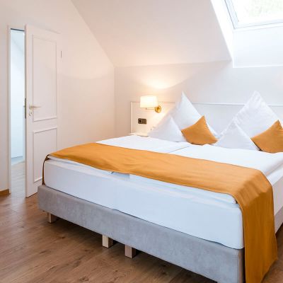 Schlafzimmer mit Boxspringbett unter einer Schräge, in der Dachschräge Fenster, links und rechts vom Bett Lampen und Ablageflächen, an der Wand hängt ein Bild, eine Tür