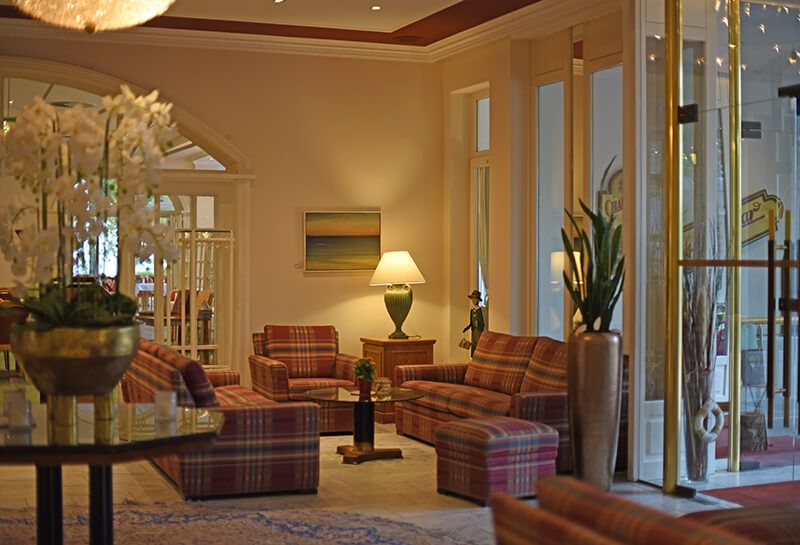 Lobby Hotel Hohe Wacht, Sitzecke mit karierten Sofas und Sesseln sowie einem Glastisch zum Verweilen, in der Ecke Tischen mit leuchtender Lampe, rechts von der Sitzecke Glastür Durchgang Kurpark, hinter der Sitzecke Durchgang zum Parkrestaurant