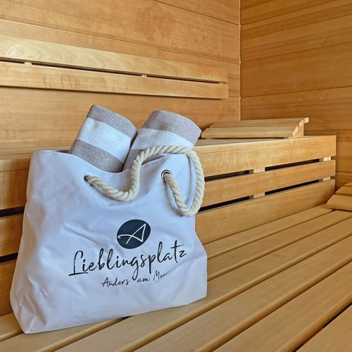 Tageswellness „Genuss am Meer“ Hohe Wacht, Sauna, weiße Wellnesstasche mit Kordelhänkel, auf der Tasche Logo Aners am Meer und Aufdruck Lieblingsplatz, aus der Tasche ragen zwei weiß-beige gestreifte eingerollte Handtücher, Tache steht auf der Holzbank