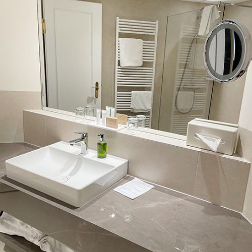 Bad im Turmzimmer mit Waschbecken, Seifenspender, Kosmetikspiegel, Kosmetikproben, Handtüchern, im Spiegel spiegelt sich Dusche und Handtuchheizkörper sowie die Tür