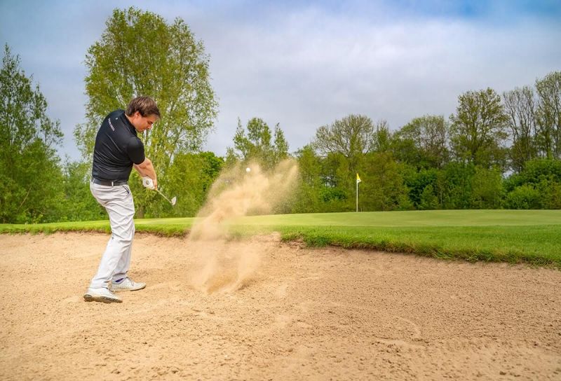 Golfplatz Hohwachter Bucht, Neudorfer Platz, im Vordergrund Golfer versucht Ball aus Sandfläche zu schlagen, aufgewirbelter Sand, vor dem Golfer das Grün mit Loch und Fähnchen, im Hintergrund Baumwand