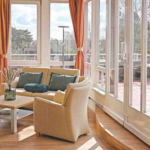 Ecke Wohnbereich Suite mit Couch, Sessel und Tisch, rundherum Fensterfront, Blick nach draussen auf den Balkon, Blick auf den Kurpark und die Ostsee