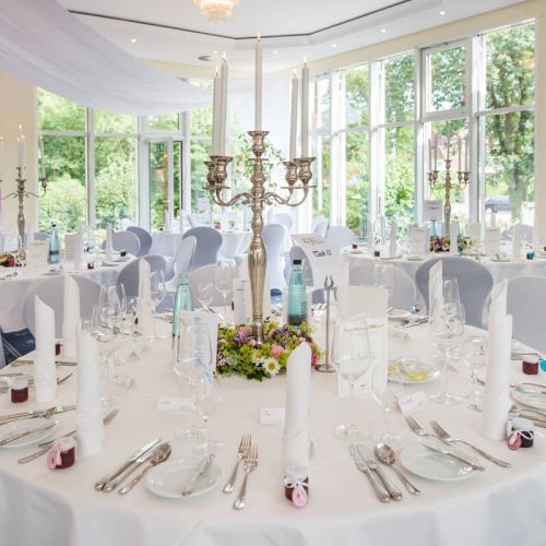 eingedeckter Banketttisch mit Geschirr, Besteck, Gläsern, Servietten, Speise- und Getränkekarte, Mitte des Tisches silberner Kronleuchter mit leuchtenden Stabkerzen und Blumendeko, im Hintergrund an der Fensterfront zwei weitere gedeckte Tische