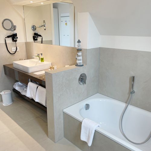 Waschbecken mit Seifenspender, Kosmetikproben, Föhn, Kosmetikspiegel, Handtücher und Kosmetikeimer, Spiegelfront, hinter einer Schamwand Badewanne, über dem Wannenrand hängt ein Handtuch