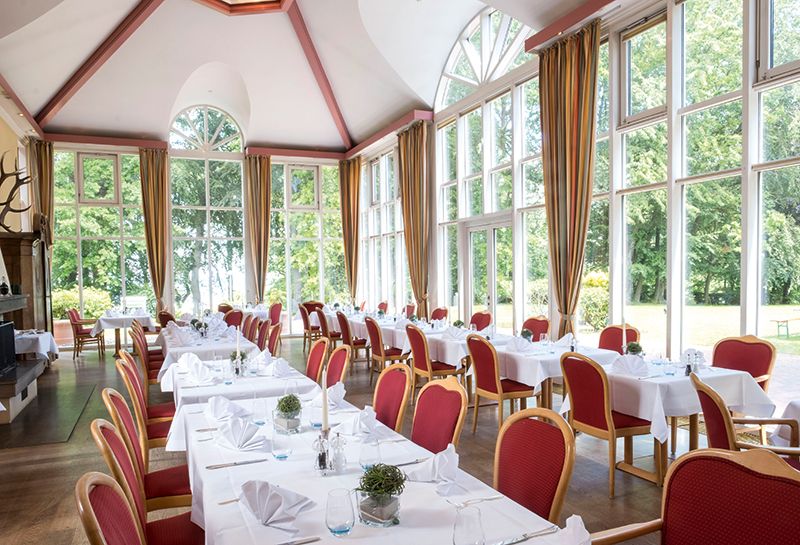 Feierlichkeiten Wintergarten, gedeckte Tische mit Betseck, Kerzen, Servietten, rote gepolsterte Stühle, Blick zur Fensterfront auf den Kurpark und die Ostsee