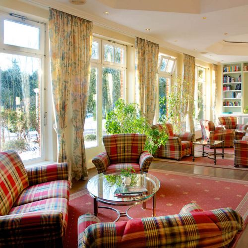 Bibliothek Hotel Hohe Wacht, verschiedene Sitzecken an der Fensterfront, karierte Sofas und Sessel mit Glastischen, zwischen den Sitzecken grüne Pflanzen, im Hintergrund sind Regale mit Büchern, Blick in den Kurpark