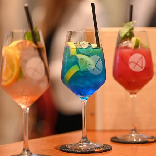 Hotelbar Hohe Wacht, auf dem Tresen drei verschiedene angemixte Cocktails im Glas auf Untersetzern, alle Gläser mit Logo Anders am Meer, Cocktails garniert mit Limetten- und Zitronenstücken, frischer Pfefferminze, Eiswürfel und schwarzen Papiertrinkhalmen
