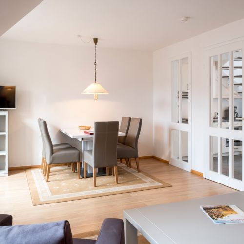 2,5-Zimmer-Ferienwohnung Essbereich, ein Tisch an der Wand, drumherum 5 Stühle, über dem Tisch hängt eine Lampe, rechts vom Tisch Zugang zur Küche, Türen mit Glaselementen, im Vordergrund Sitzecke angeschnitten