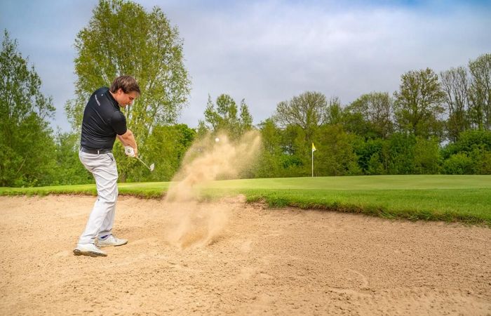 Golfplatz Hohwachter Bucht, Neudorfer Platz, im Vordergrund Golfer versucht Ball aus Sandfläche zu schlagen, aufgewirbelter Sand, vor dem Golfer das Grün mit Loch und Fähnchen, im Hintergrund Baumwand