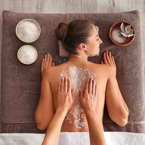 Wellness und Spa im Hotel Hohe Wacht, Frau mit freiem Oberkörper auf dem Bauch liegend auf Massageliege, Haare zu einem Dutt zusammen, Zwei Hände massieren den Rücken der Frau, Salzpeeling