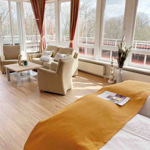 Suite mit Wohn- und Schlafbereich, Sitzecke mit Sofa, Sessel und Tisch, Doppelbett mit Tagesdecke und Prospekt, helle Fensterfront, umliegender Balkon mit Blick auf die Ostsee