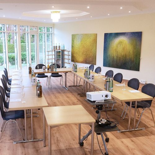 Tagungsraum Lütjenburg im Hotel Hohe Wacht, Tische in Stühle in U-Form aufgestellt, eingedeckt mit Gläsern, Getränken und Schreibblöcken, im Vordergrund Beamer, Kronleuchter an Decke, Fensterfront, an der rechten Wand hängen zwei große gemalte Bilder