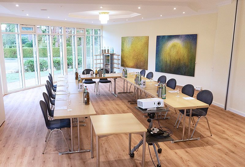 Tagungsraum Lütjenburg im Hotel Hohe Wacht, Bestuhlung in U-Form, Tische eingedeckt mit Gläsern, Getränken und Schreibblöcken, im Vordergrund Beamer, Kronleuchter an Decke, Fensterfront, an der rechten Wand hängen zwei große gemalte Bilder