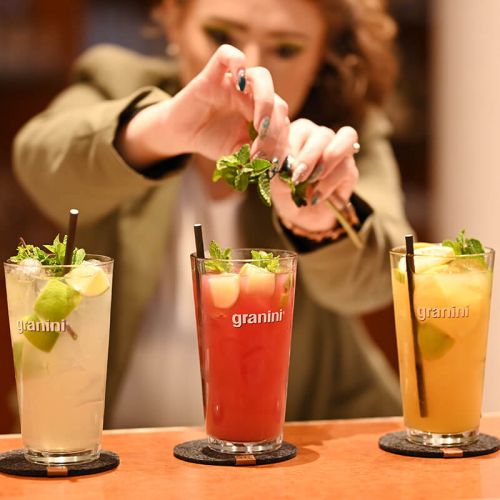 Getränkekarte Parkrestaurant Hohe Wacht, Frau hinterm Tresen garniert die befüllten Gläser im Vordergrund mit frischer Minze, in den Gläsern sind verschiedene Granini Limonaden mit geschnittener Limette, Eiswürfeln und schwarzem Papiertrinkhalm