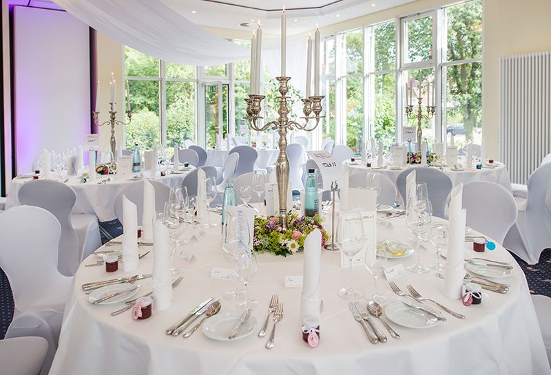 eingedeckter Banketttisch mit Geschirr, Besteck, Gläsern, Servietten, Speise- und Getränkekarte, Mitte des Tisches silberner Kronleuchter mit leuchtenden Stabkerzen und Blumendeko, im Hintergrund an der Fensterfront zwei weitere gedeckte Tische