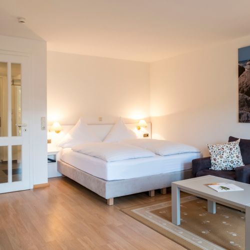 1-Zimmer-Ferienwohnung Schlafbereich mit Boxspringbett, links und rechts neben dem Bett Lampen und Ablageflächen, links Eingangstür, rechts Sitzecke angeschnitten