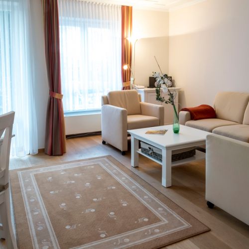 2-Zimmer-Komfortappartement mit Sitzecke am Fenster, Sofa mit roter Decke, zwei Sessel und Tisch, in der Ecke Sideboard mit Radio und Boxen