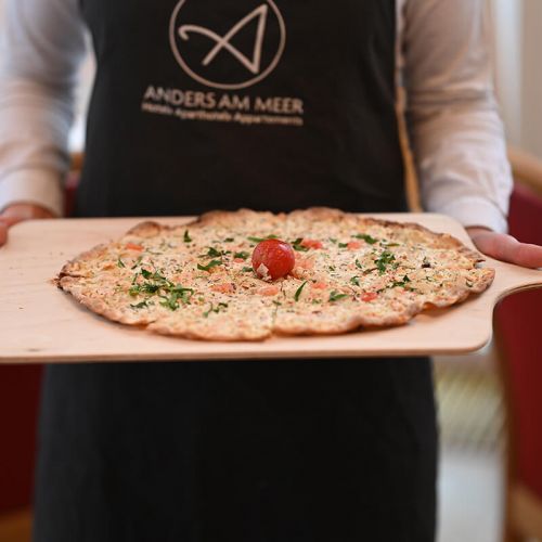 Speisekarte Parkrestaurant Hohe Wacht, Bedienung im Hintergrund mit schwarzer Schürze mit Anders am Meer Logo, im Vordergrund häält die Person eine Holzbrett mit einem Lachs Flammkauchen