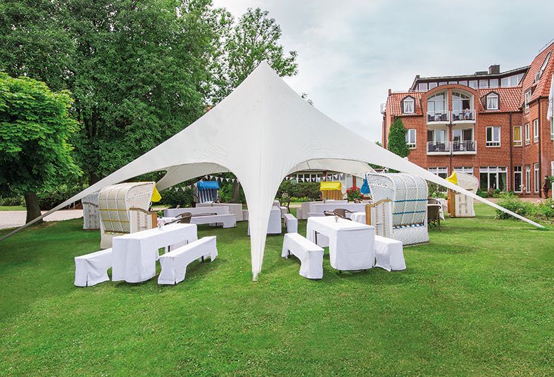 Kurpark Hotel Hohe Wacht, offener Pavillion, unterm Pavillion weiß bezogene Tische und Bänke, Strandkörbe als Sitzgelenheiten, Wimpelgirlanden, im Hiontergrund Rückansicht Hotel Hohe Wacht