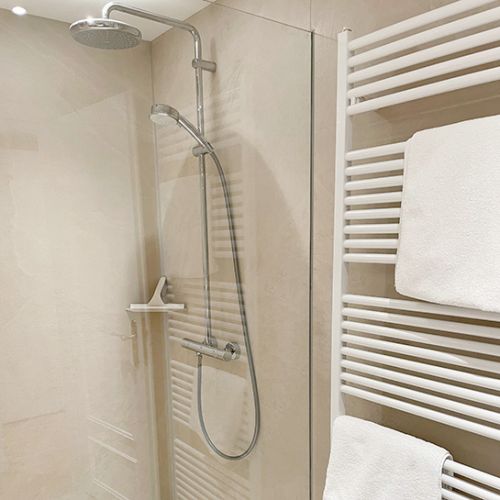 Dusche mit Glaswänden, durch die Glaswand sieht man die Armatur mit großer und kleiner Duschbrause, rechts neben der Dusche hängt der Handtuchwandtrockner mit weißen Handtüchern