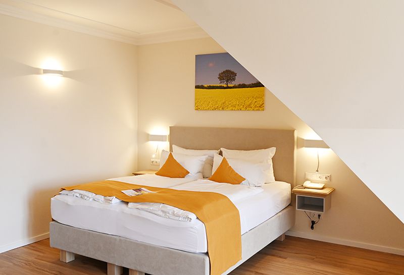 Boxspringbett unter der Schräge, über dem Kopfende Leinwand mit Rapsmotiv, links und rechts vom Bett Lampen und Ablageflächen