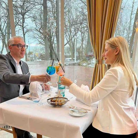 Pärchen im Parkrestaurant Hotel Hohe Wacht am Fenster, das Pärschen stößt mit Cocktails an, auf dem Tisch Teller, Besteck, Gläser, Wasserflasche, Brotkorb und Aufstrich, im Hintergrund Blick auf Ostsee