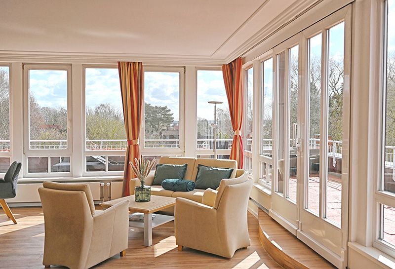 Sitzecke in der Kurpark-Suite, Sofa mit Kissen und Decke, zwei Sessel und Tisch, komplette Fensterfront, Terrassen zum Balkon rundum mit Blick in den Kurpark und auf die Ostsee