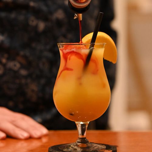 Getränkekarte Parkrestaurant Hohe Wacht, Person angeschnitten und verschwommen steht am Tresen im Hintergrund, davor Nahaufnahme Glas Cocktail auf Untersetzer, garniert mit Orangenscheibe am Glasrand, roter Likör wird reingegossen, schwarzer Papierhalm