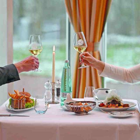 gedeckter Tisch mit fertigen Speisen im Restaurant, zwei zuprostende Hände mit Glas Weißwein, brennende Kerze in der Mitte vom Tisch