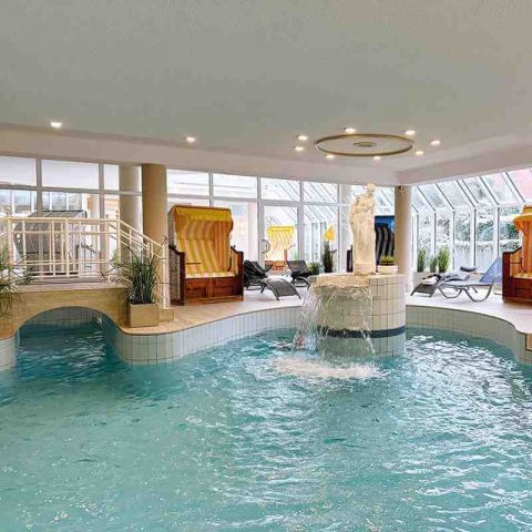 Wellness und Spa Hohe Wacht, Ausschnitt des Wellnessbereichs mit Schwimmbad, Sttrandkörbe und Liegen zum Entspannen, im Schwimmbecken Statue mit Wasserfall, kleine Brücke über dem Schwimmbecken, Glasfronten