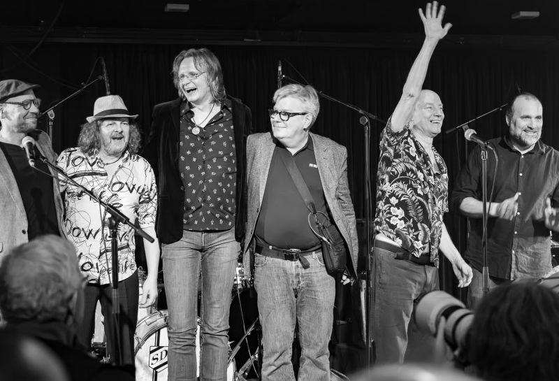 Man sieht die Band Mojo Men ist auf einer Bühne. Es sind 6 Bandmitglieder zu sehen, die ins Publikum schauen. Schwarz-Weiß-Aufnahme