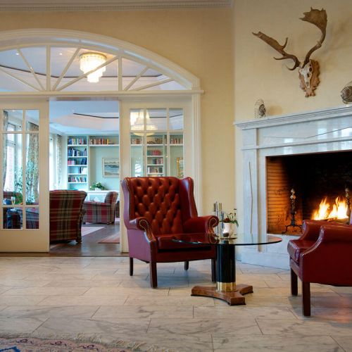 Lobby Hotel Hohe Wacht, mit Blick in die Bibliothek, vor der Bibliothek brennender Kamin mit zwei großen weinroten Ledersesseln und einem Glastisch zum Entspannen, über dem Kamin hängt ein Geweih